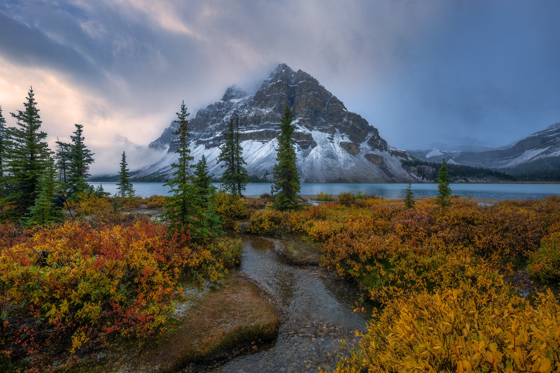Bow Lake Bow Lake in Banff National Park with golden fall foliage along the shoreline and snow-dusted mountain peaks reflected in calm, glacier-fed water.